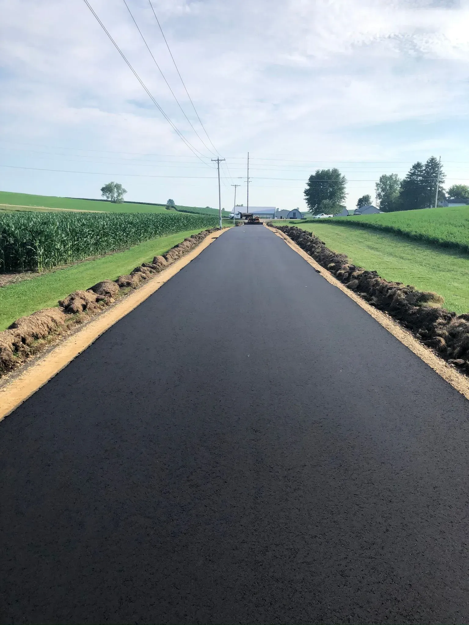 A freshly paved black asphalt road stretches into the distance, flanked by green fields and dirt shoulders. A newly paved, dark asphalt road stretches into the distance, flanked by green fields and fresh soil shoulders.