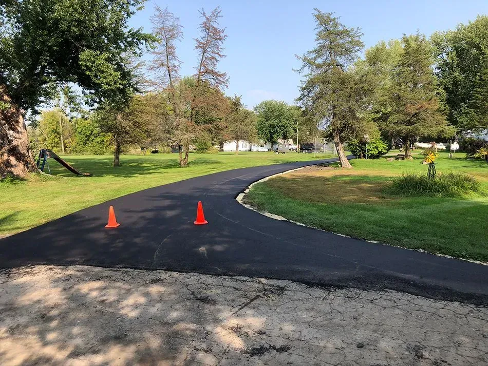 A freshly paved asphalt driveway curves through a green grassy lawn, with two orange traffic cones on the surface. A freshly paved black asphalt driveway curves through a green, grassy yard, marked by two small orange traffic cones.