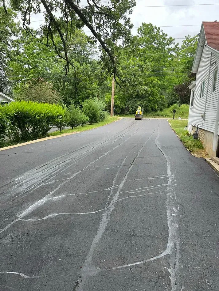 A construction roller smooths a newly paved asphalt driveway next to a white house with trees in the background. A freshly paved asphalt driveway with visible sealant lines, leading toward a house and a small road roller in the distance.