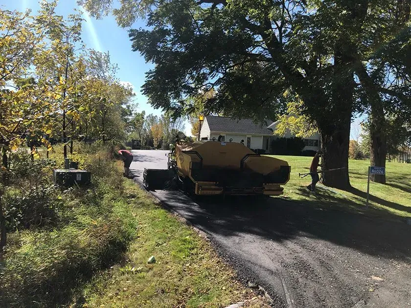 Workers operate paving equipment to resurface a driveway on a sunny, tree-lined residential property. Workers operate a yellow paving machine to resurface a driveway on a sunny day with trees in the background.