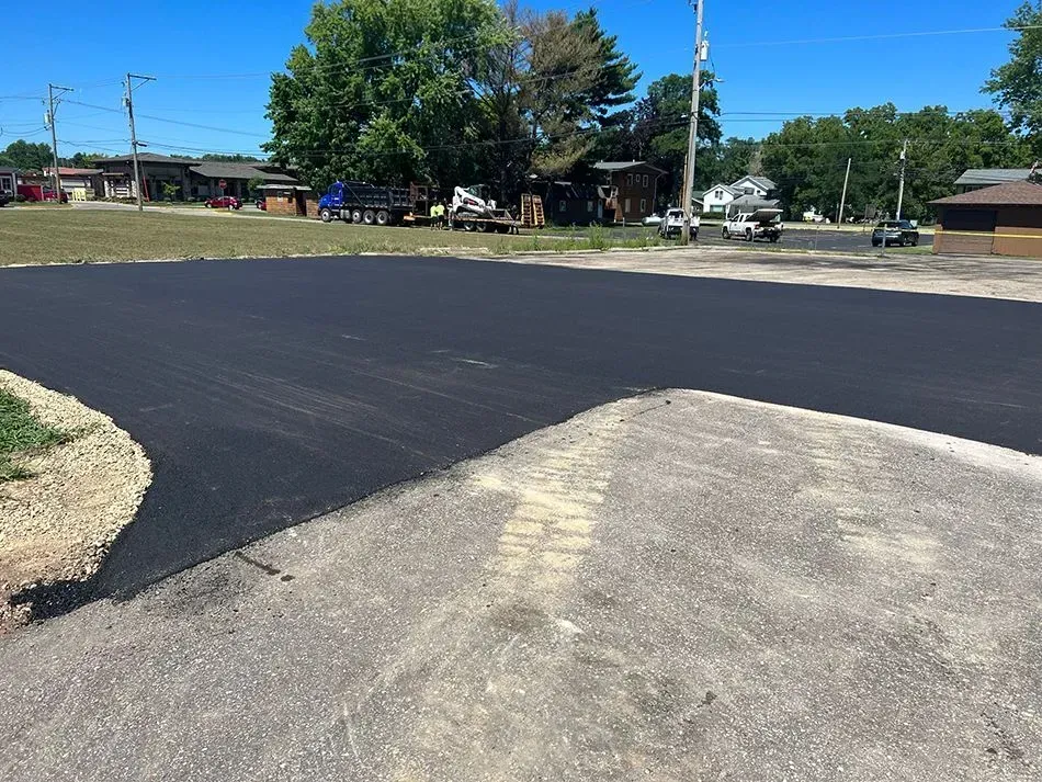 A freshly paved dark asphalt patch contrasts with an older, lighter, gravel-textured parking lot surface outdoors. A contrast between freshly laid dark black asphalt and an older, textured gray gravel surface in an outdoor parking lot.