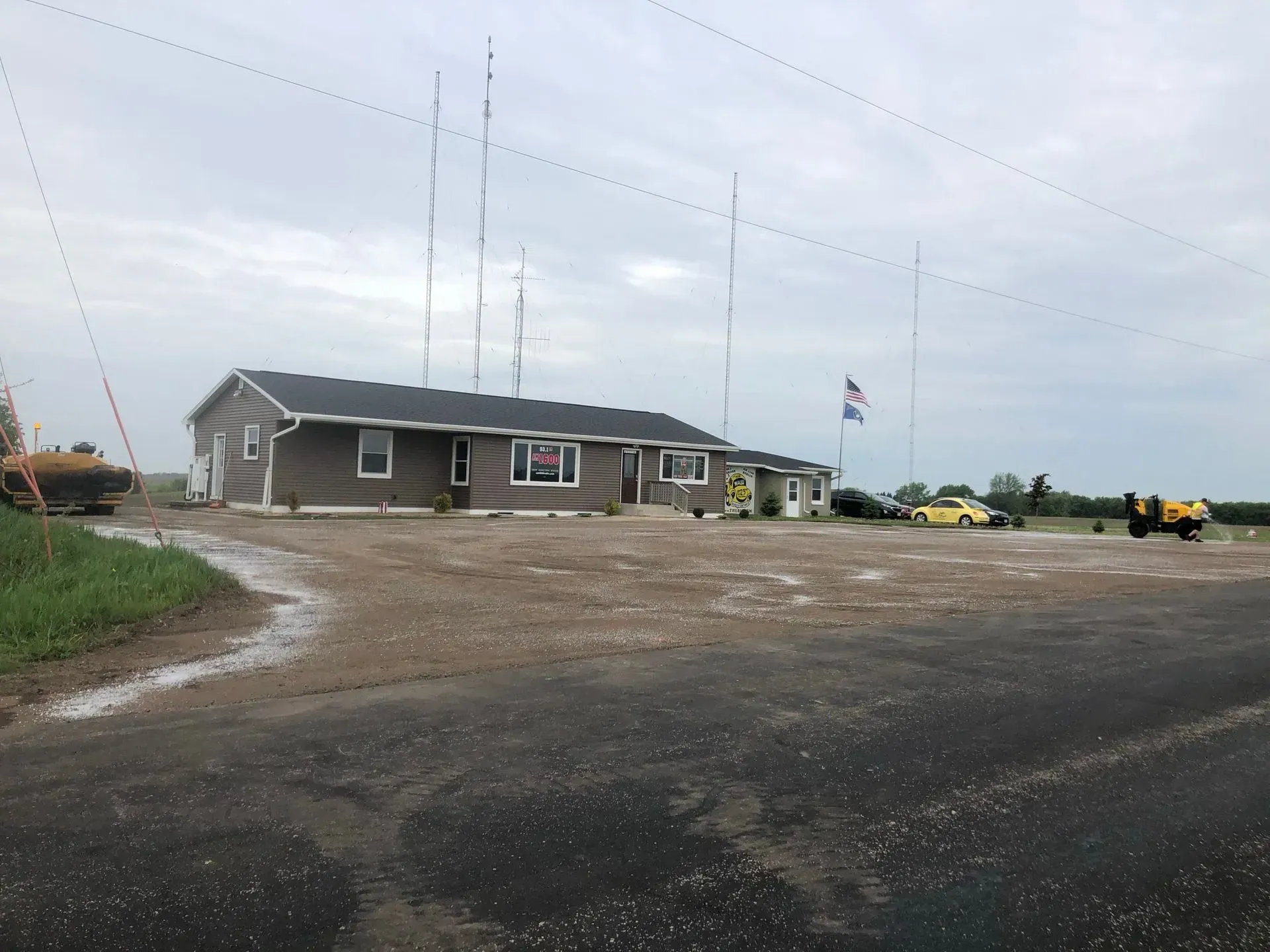 A gray building with radio towers in the background, set on a gravel lot next to an asphalt road under a cloudy sky. A gray, single-story building with tall antenna towers, a flagpole, and a gravel parking lot under a cloudy sky.