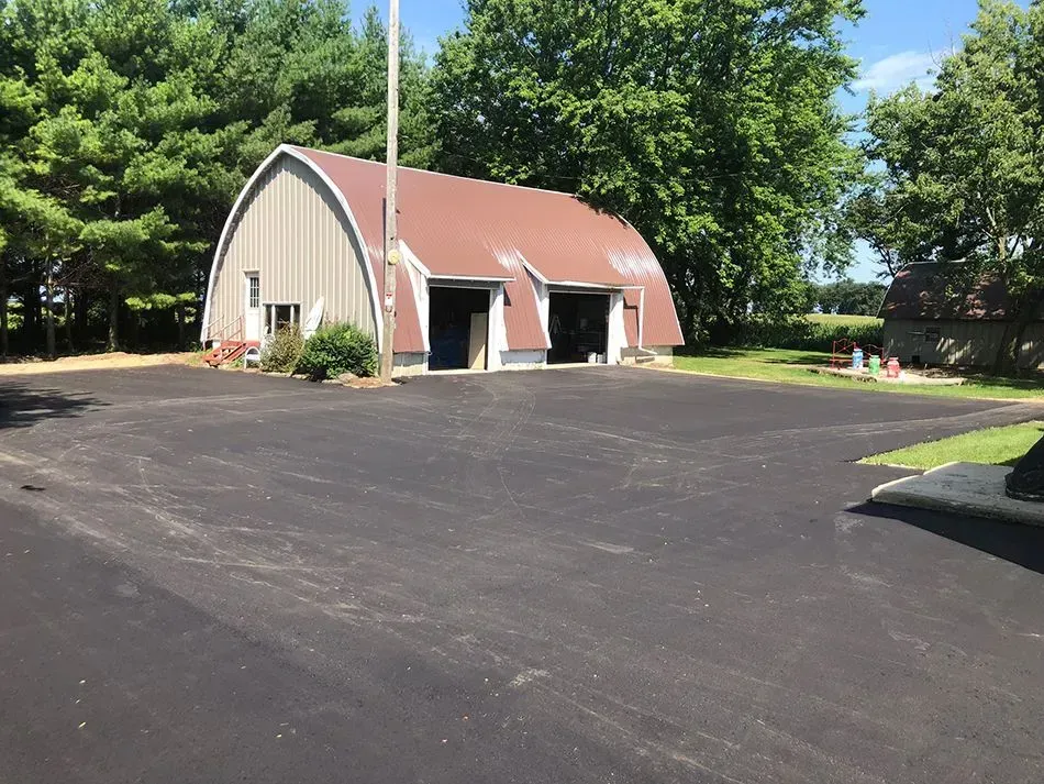 A paved driveway leads to a Quonset-style building with a reddish roof, surrounded by trees on a sunny day. A tan, arched-roof building with two garage bays sits beside a large, freshly paved black asphalt parking lot.