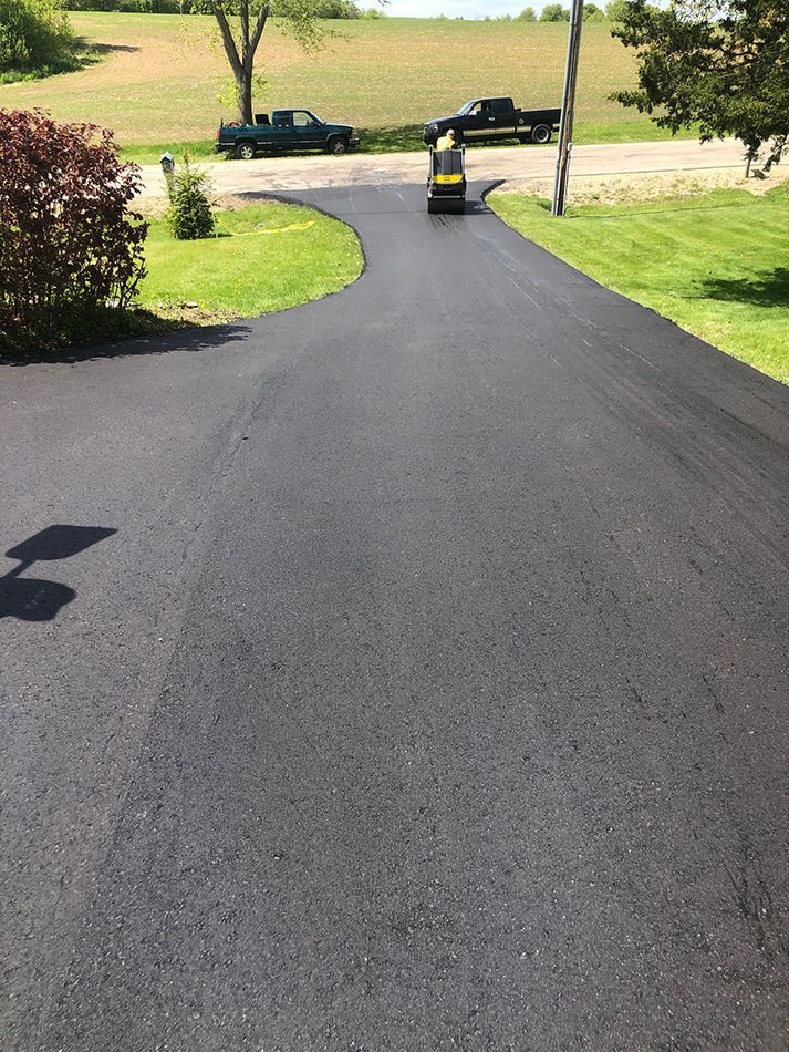 A freshly paved black asphalt driveway curves towards a road with two parked trucks in the background. A freshly paved asphalt driveway curves toward a road with two parked pickup trucks in the background.