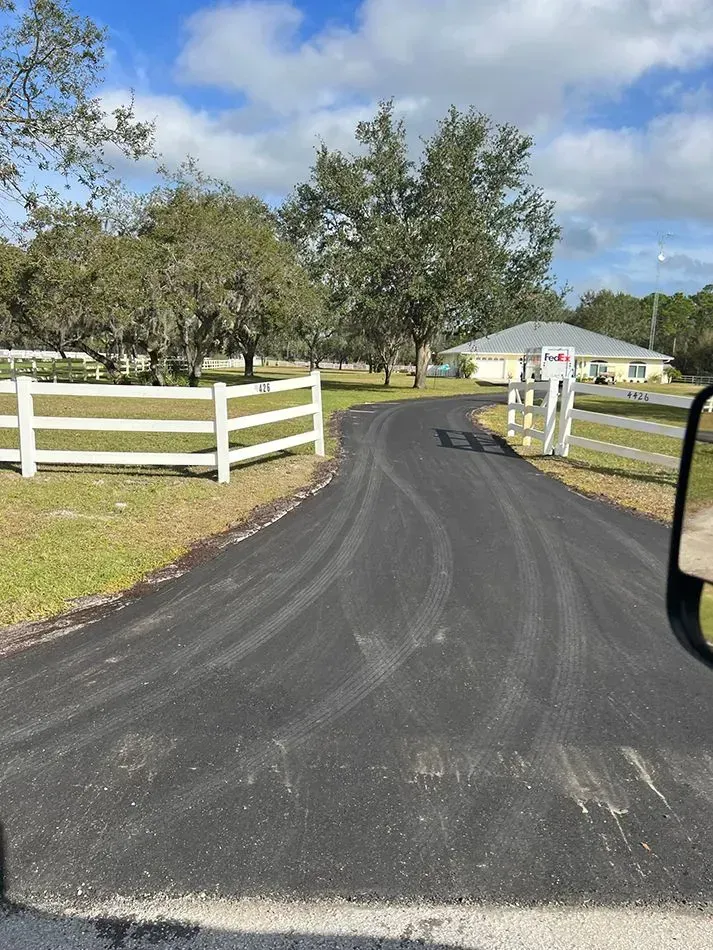 A paved driveway winds through a grassy yard toward a house in the distance, framed by white fences and mature trees. A black paved driveway leading to a house in the distance, framed by white wooden fences and trees under a blue sky.
