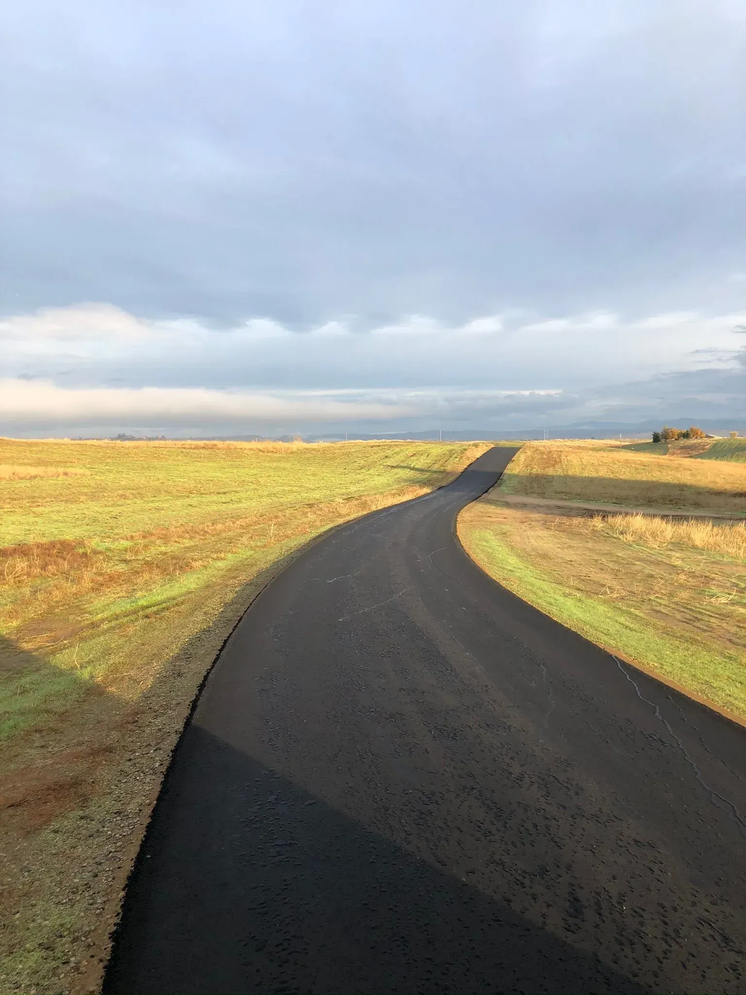 A freshly paved, winding asphalt path cuts through a grassy field under a cloudy, overcast sky. A paved asphalt path winds through a grassy field under a cloudy sky.