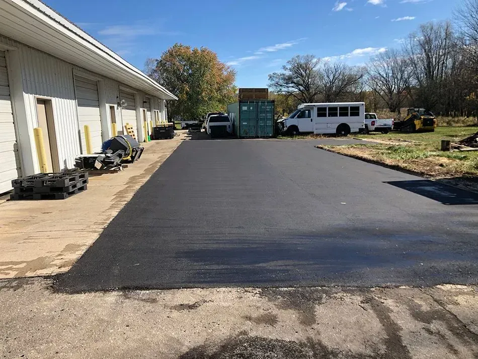 A newly paved dark asphalt driveway leads past a commercial metal building toward a shipping container and a parked bus. A newly paved asphalt driveway next to a warehouse building, with a white shuttle bus and storage container in the back.