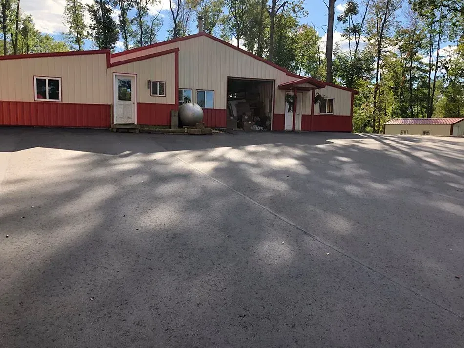 A beige and red metal warehouse with an open bay door, a side entrance, and a large propane tank on a sunny paved lot. A wide view of a beige and red metal building with a large open garage door and a paved parking area, surrounded by trees.