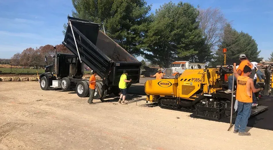 A dump truck empties material into a yellow asphalt paver while road workers operate the machinery on a gravel site. A dump truck is discharging material into a yellow asphalt paver while road workers operate the machinery outdoors.