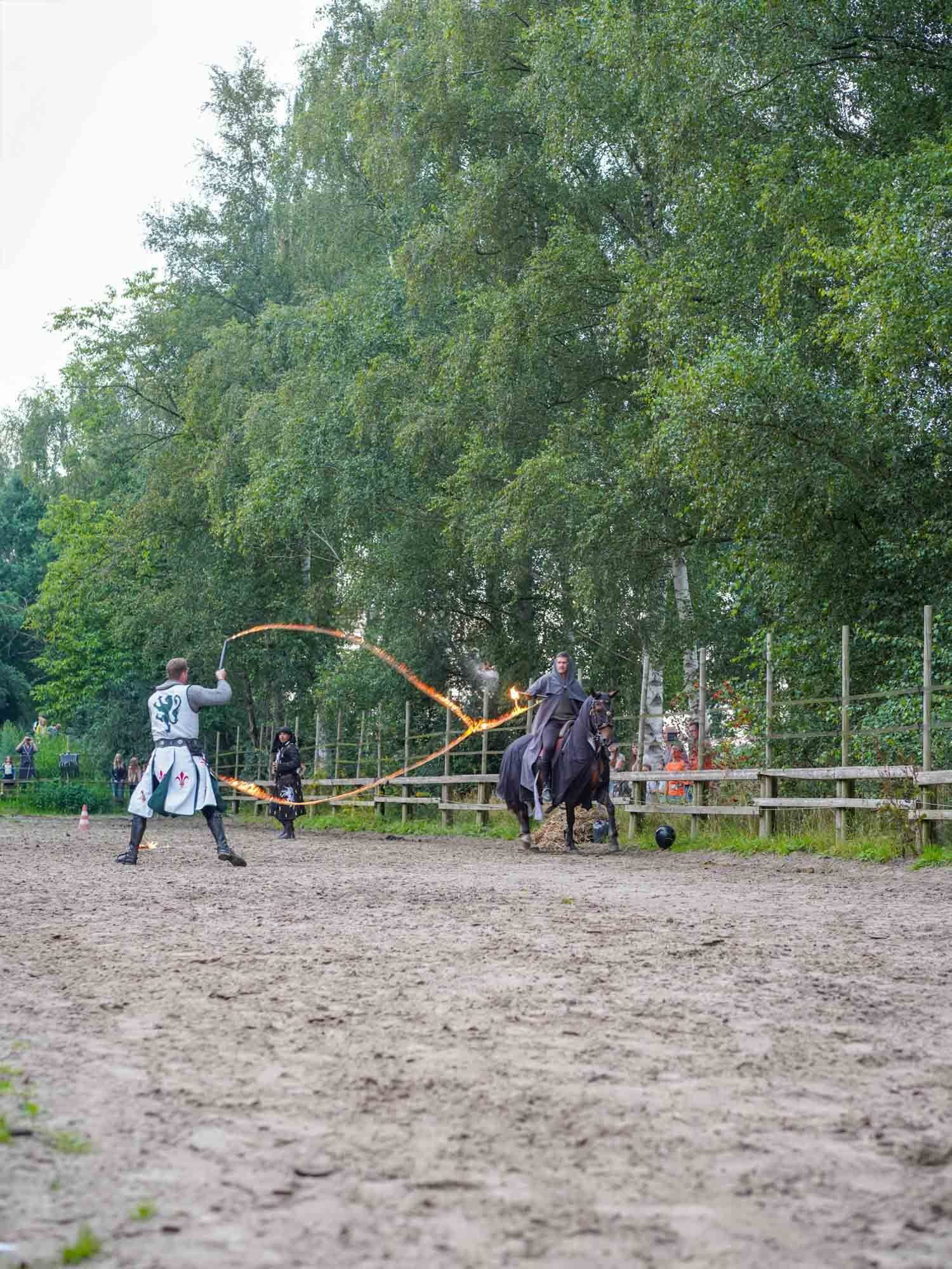 Zwei Reiter üben auf dem Pferd das Lassowerfen in einer matschigen Arena, im Hintergrund sind Bäume zu sehen