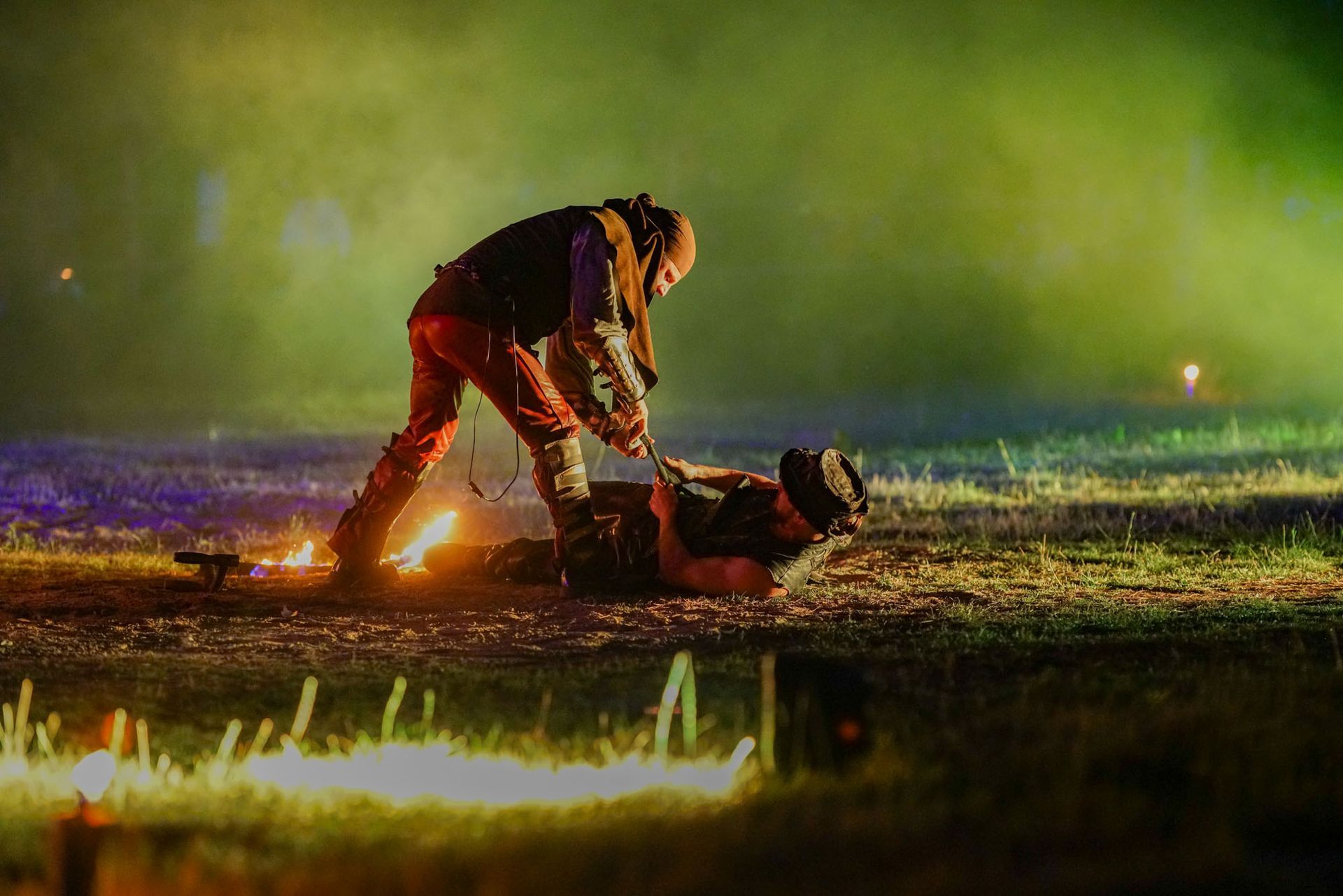 Eine Person beugt sich nachts über eine andere, die neben einem kleinen Feuer im Gras liegt. Funken steigen auf, im Hintergrund ist es grün