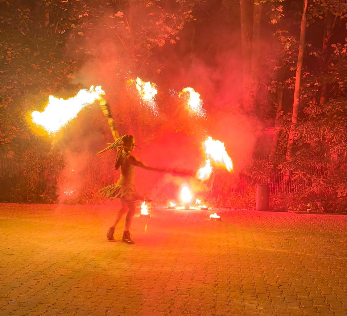 Eine Person wirbelt brennende Stäbe auf einer Waldlichtung, umgeben von rotem Rauch und Feuer.