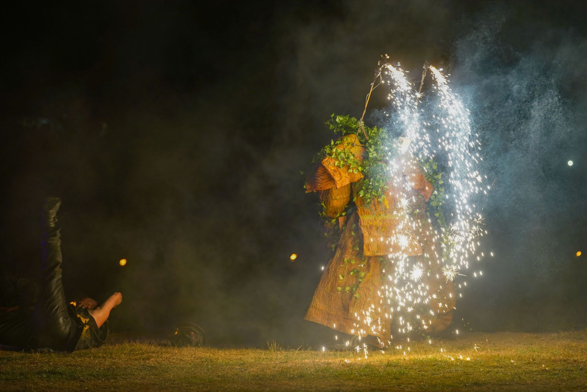 Eine verkleidete Person läuft nachts vor einem farbenfrohen Feuerwerk, umgeben von Rauch und Funken bei einer Show.