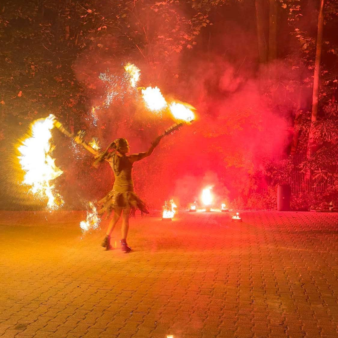 Frau im Kleid führt in einer rötlichen Umgebung im Freien einen Feuertanz mit Flammen auf.