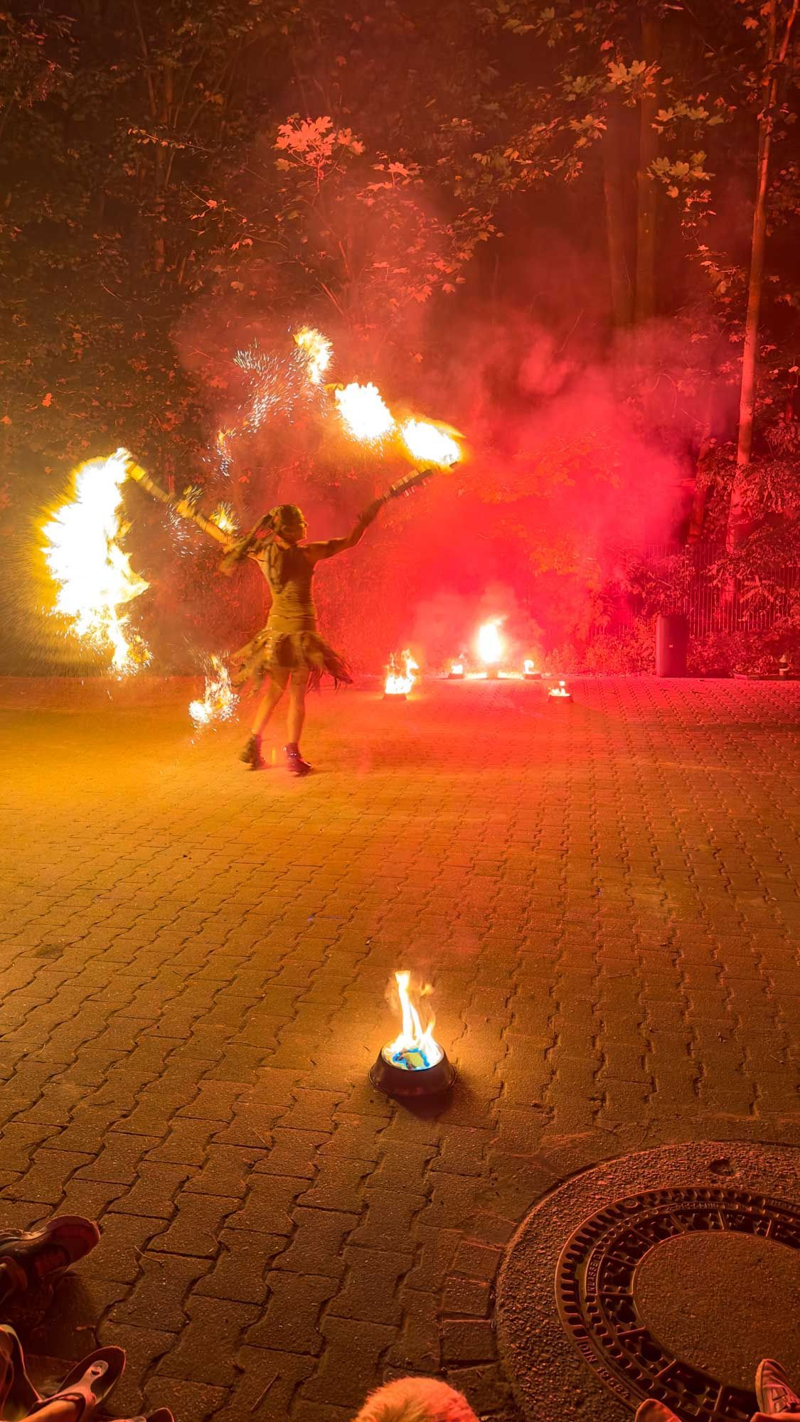 Eine Person führt nachts einen Feuertanz auf und ist auf einer gepflasterten Fläche von leuchtend roten und orangefarbenen Flammen umgeben.