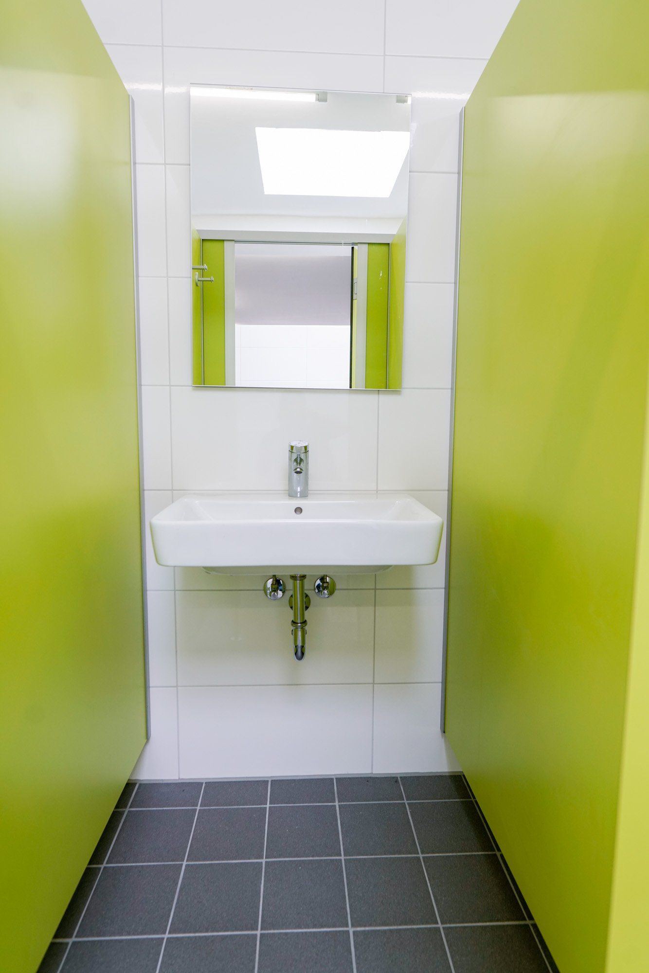 Hellgrüne Kabinentüren rahmen ein weißes Waschbecken und einen Spiegel in einer öffentlichen Toilette ein.