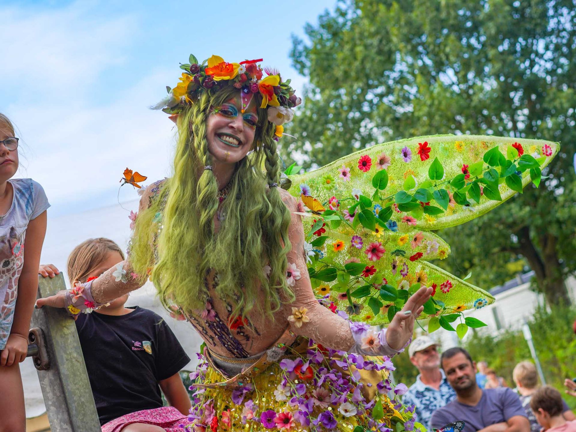 Kostümierte als Elfe verkleidete Person mit Blumenkranz und grünen Flügeln lächelt in die Kamera im Rahmen der Stelzenperformance