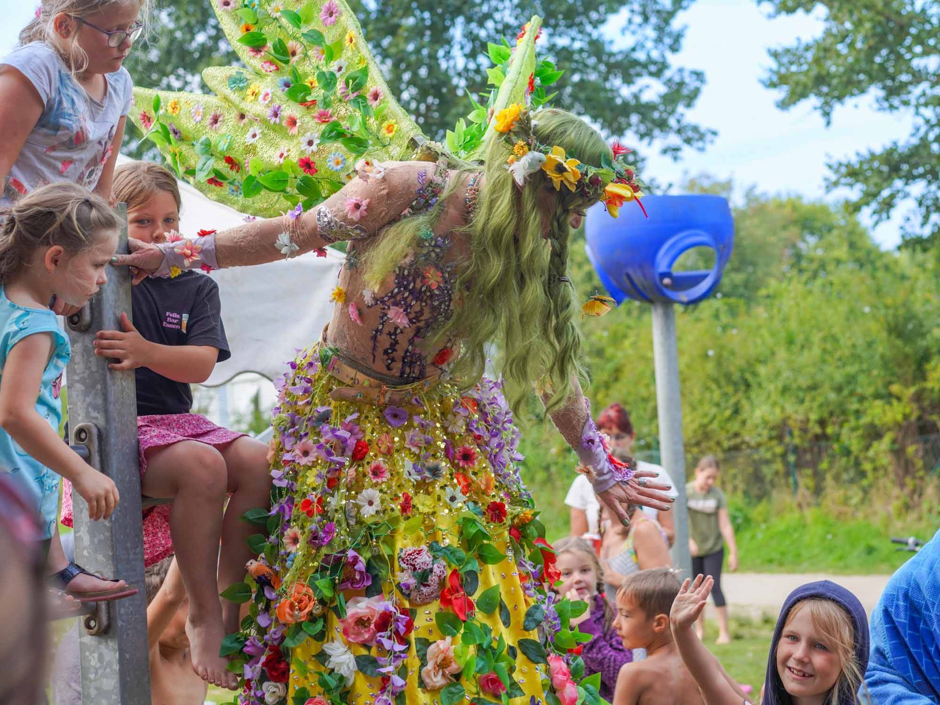 Fee als Stelzenläuferin im Blumenkostüm interagiert mit Kindern 