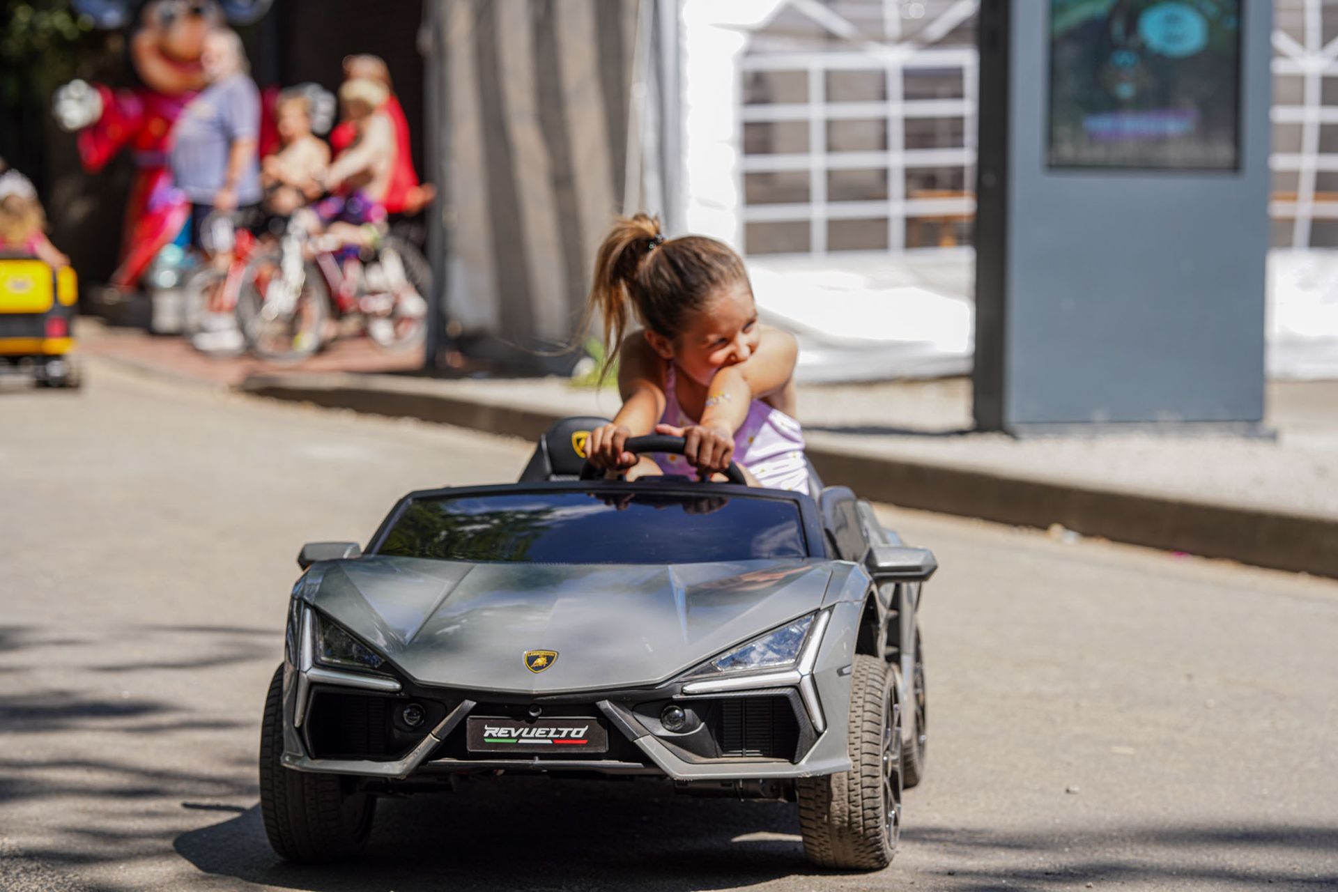 Ein Kind fährt auf einer Straße ein schwarzes Spielzeug-Sportauto, während die Fußgänger im Hintergrund unscharf sind