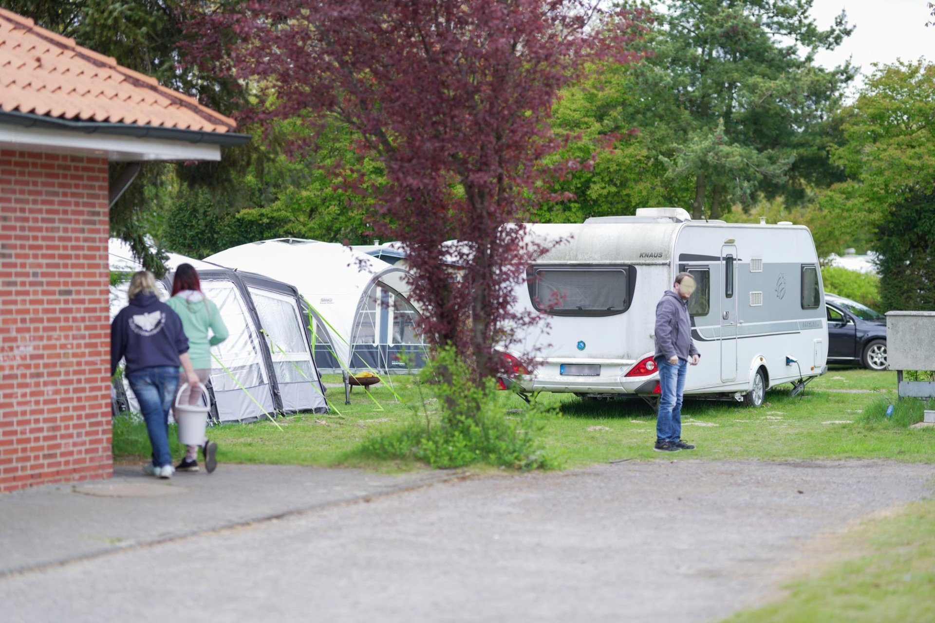 Menschen in der Nähe eines Wohnmobils und eines Zeltes auf einem Campingplatz. Einer trägt einen Eimer. 