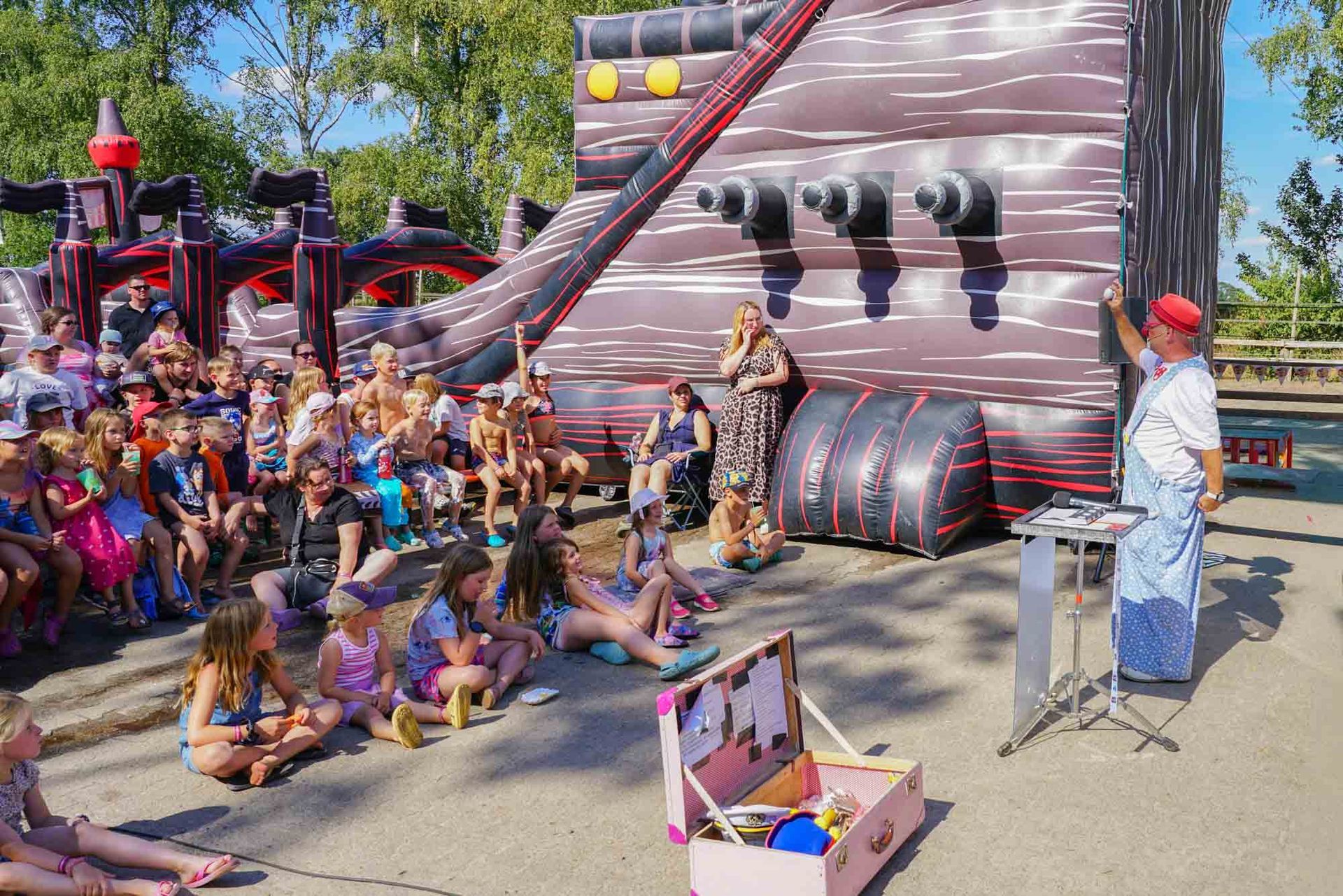 Clown Maxxx Show im Freien mit Kindern, die auf dem Boden sitzen und einer Vorführung neben einer gestreiften Hüpfburg zusehen.