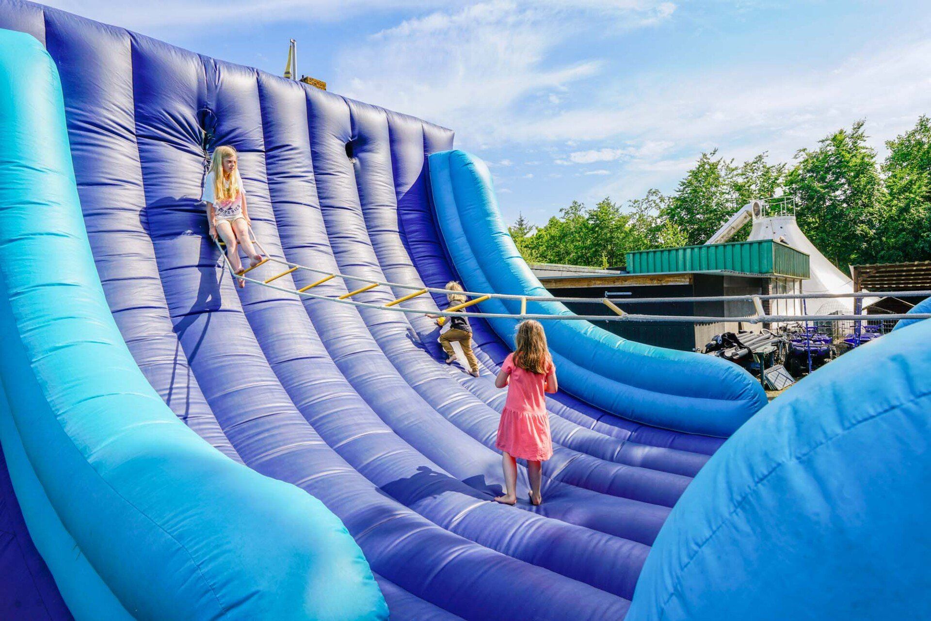 Kinder spielen an einem sonnigen Tag auf einer riesigen blauen aufblasbaren Rutsche mit Seilen.