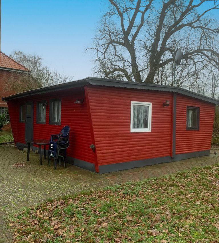 Rote Hütte mit dunkler Verzierung, kleiner Tisch und Stühle draußen. Grau gepflasterter Bereich, grünes Gras und kahle Bäume.