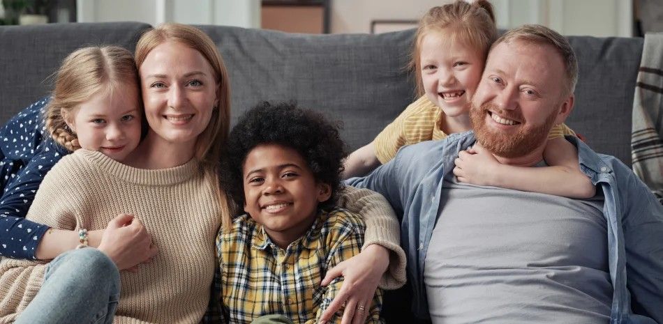 Five people smiling on a couch in a cozy living room, with two children and three adults close together.