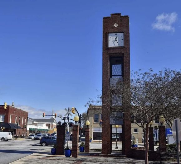 Brick clock tower in a town square under a blue sky, with trees, benches, and parked cars nearby