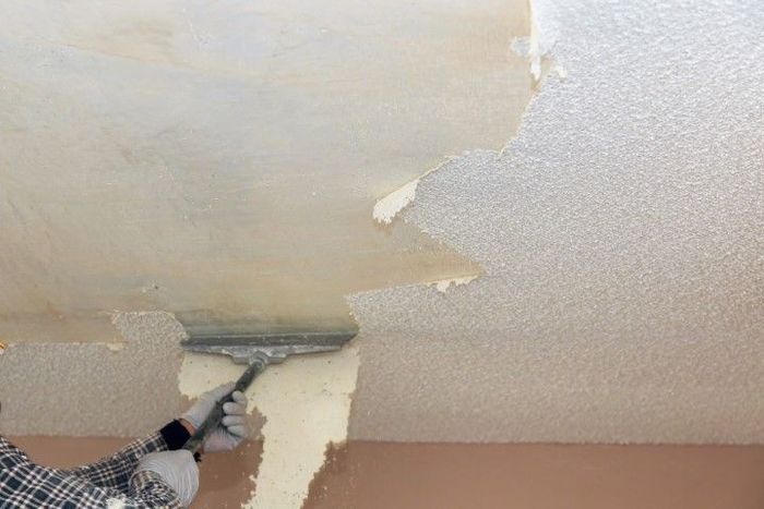 Person scraping textured ceiling with a wide scraper, removing old paint.