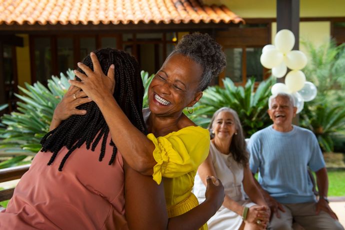 Two people hugging outdoors, with smiling guests seated nearby under white balloons.
