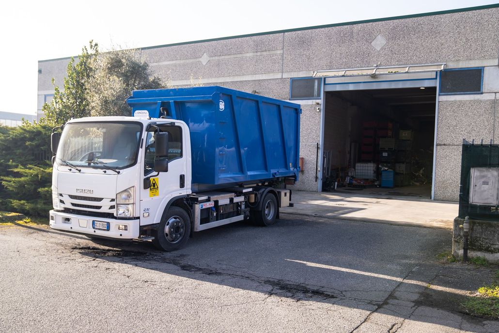 White Isuzu truck with blue dumpster in front of a building with open garage door.