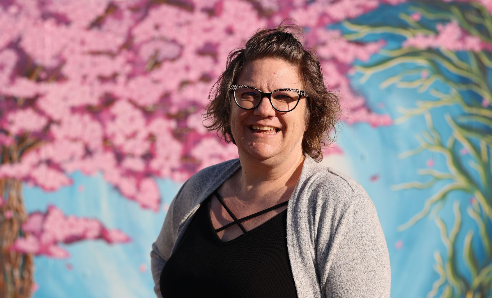 Smiling person in glasses and gray cardigan standing before a pink-and-blue mural of blossoms and vines