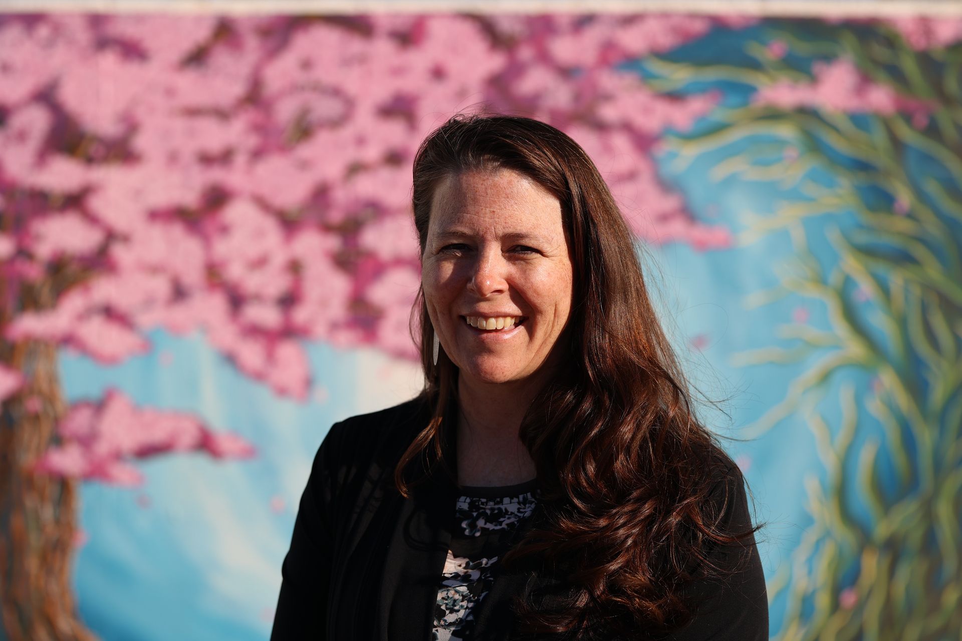 Smiling woman in a black jacket standing before a colorful mural with pink blossoms and blue leaves