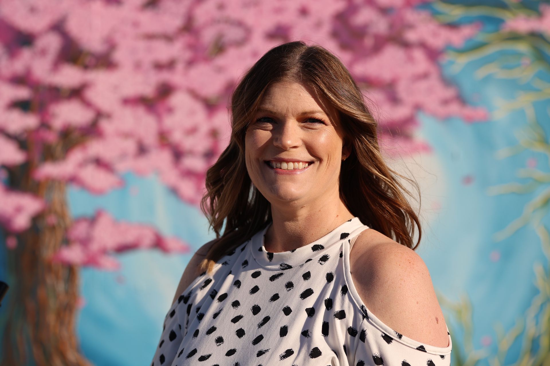 Woman smiling in a polka-dot blouse, standing before a pink blossom mural.