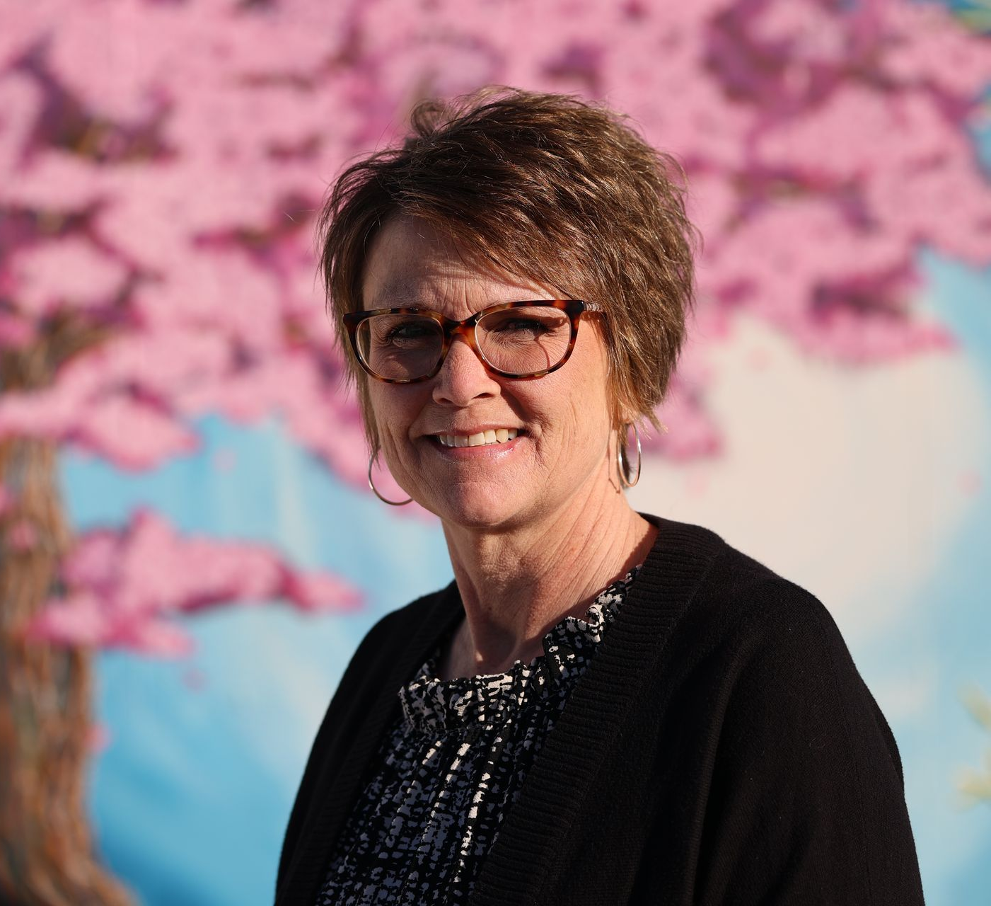 Smiling woman in glasses and black cardigan before a pink blossom backdrop