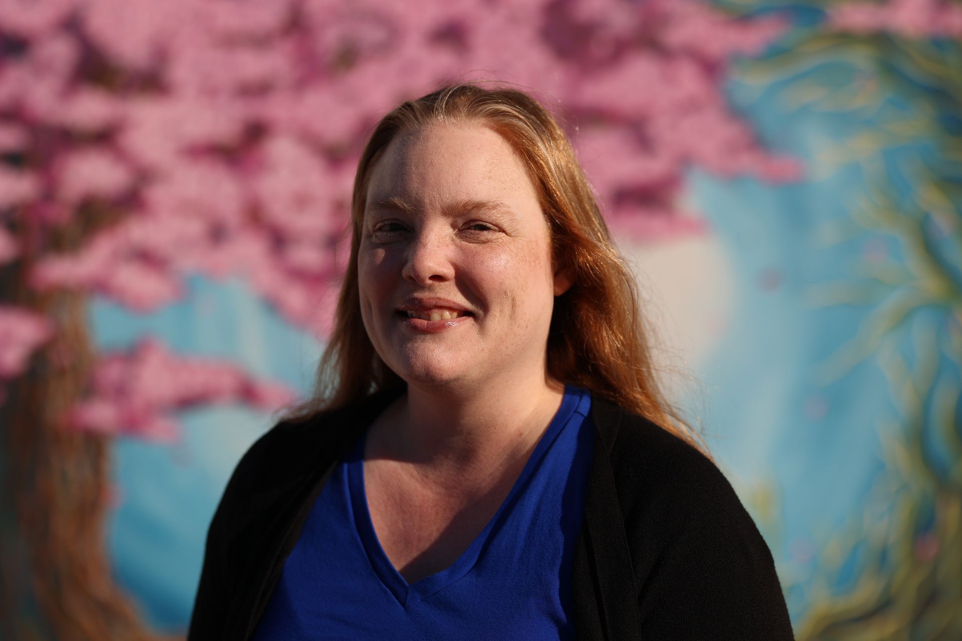 Smiling woman in a blue shirt and black cardigan, standing before a pink-blossomed mural.