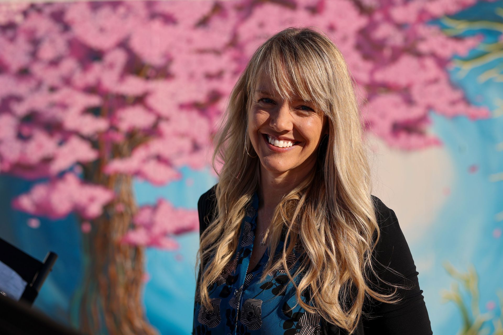 Smiling blonde person in a dark jacket against pink blossoms and a blue mural background