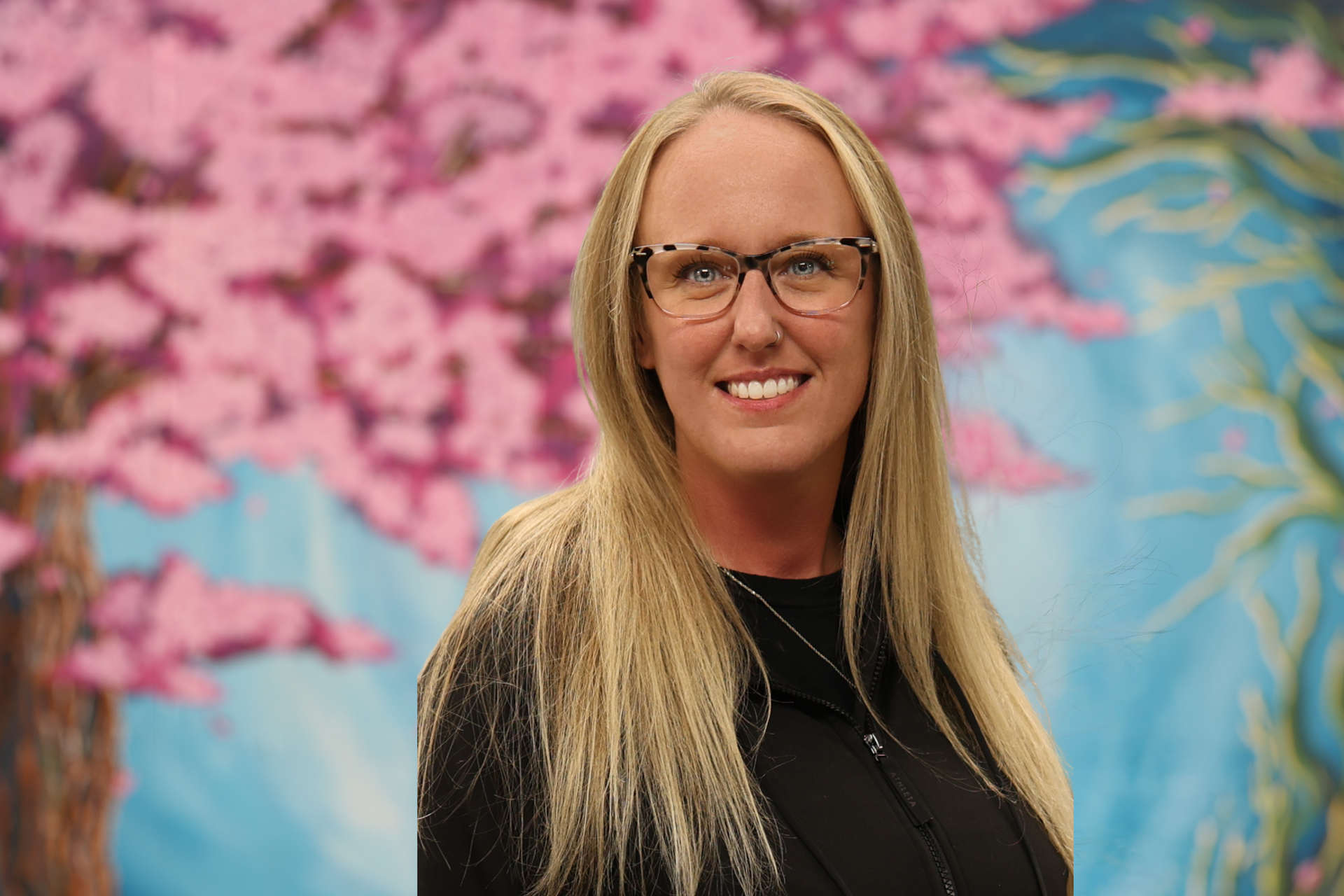 Woman with long blonde hair and glasses smiling in front of a colorful floral mural