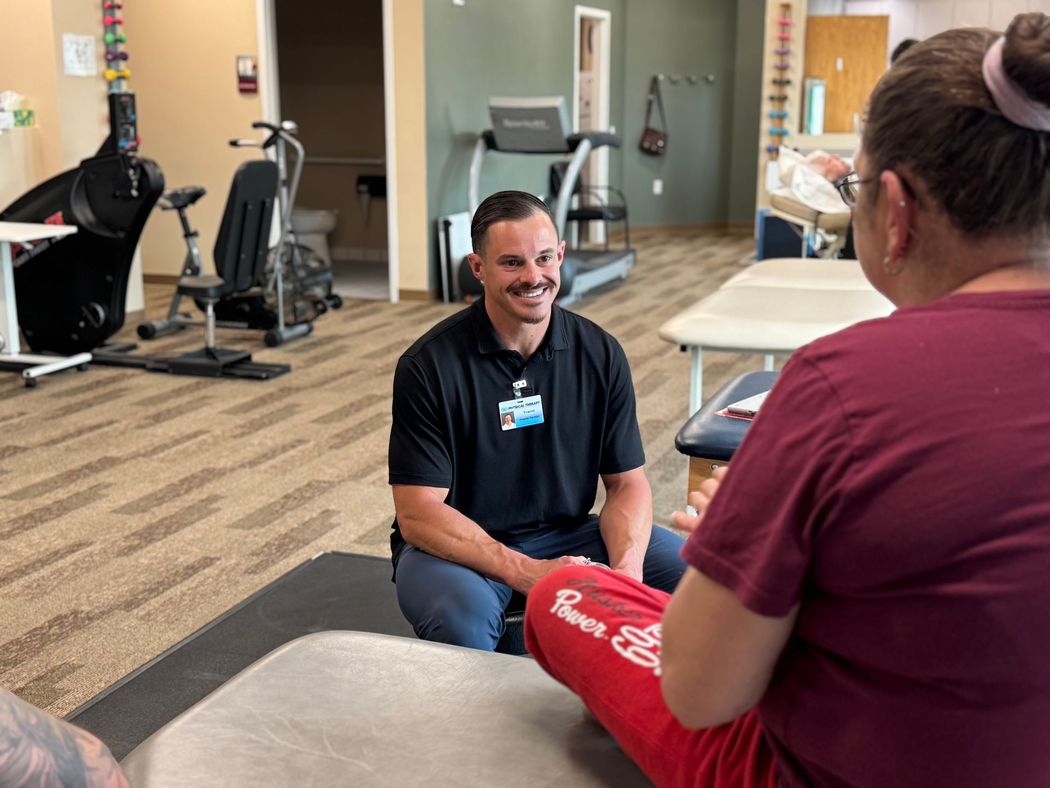 Two people talking in a gym or rehab room, one kneeling and smiling while the other sits nearby.