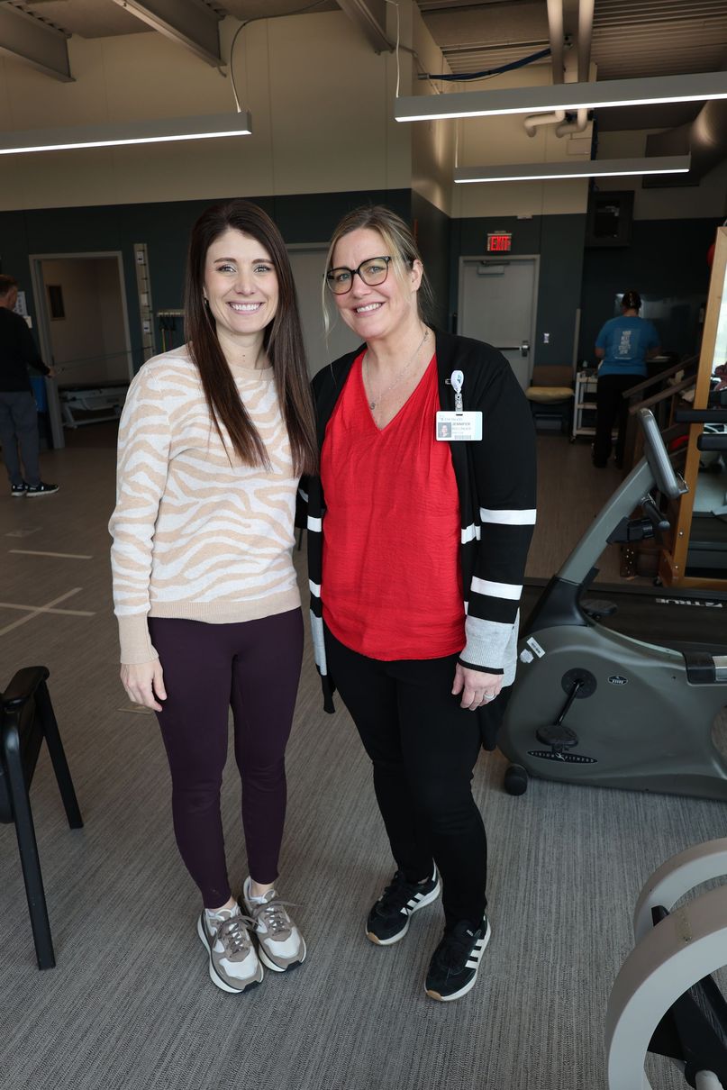 Two women standing together in a gym, smiling, with exercise machines in the background.