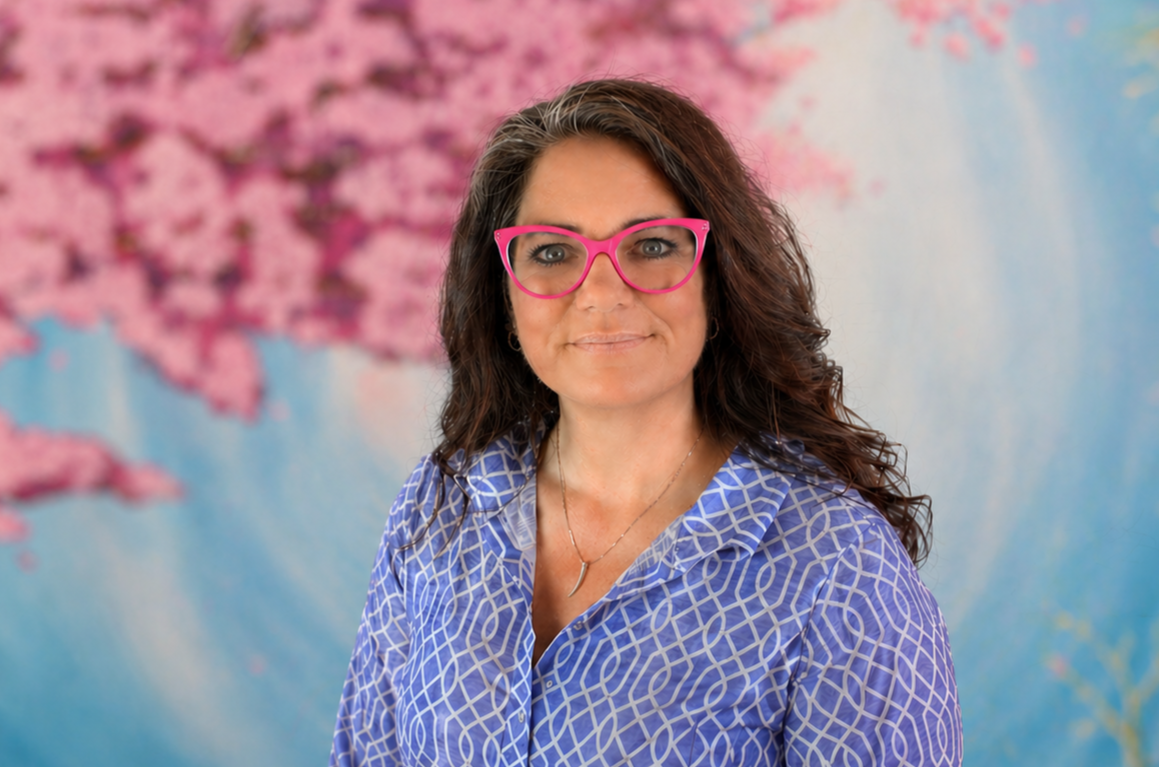 Woman in pink glasses and blue patterned blouse, smiling against a soft floral backdrop