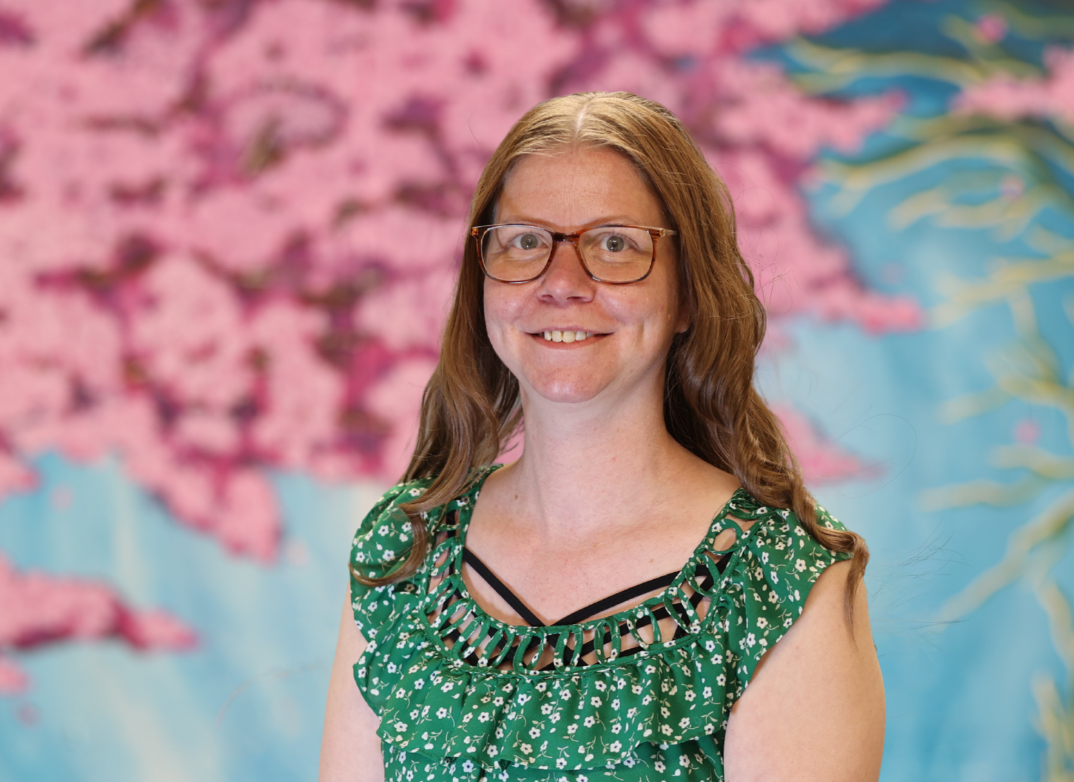 Woman in glasses wearing a green patterned top, smiling against a pink and blue mural background