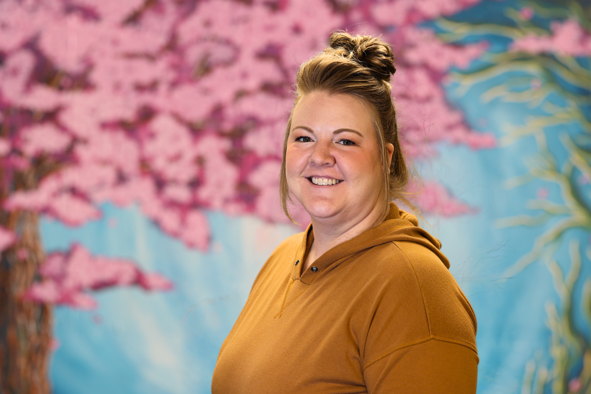Smiling person in a mustard hoodie against a colorful floral mural background