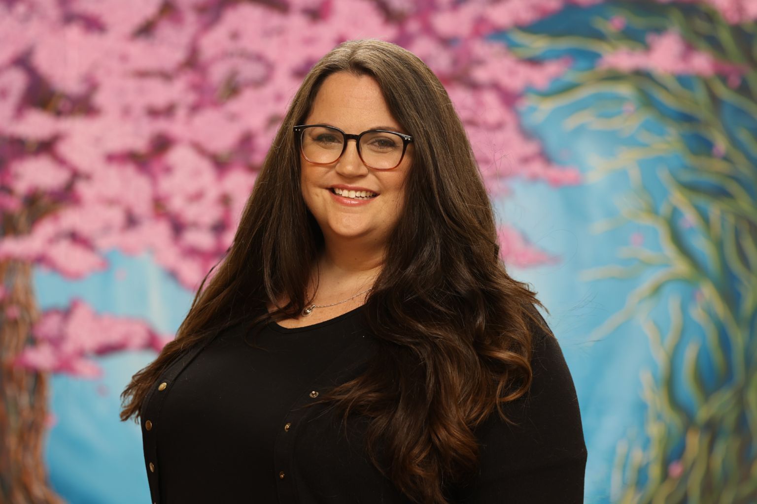 Smiling person in glasses and brown sweater against a blue and pink floral backdrop