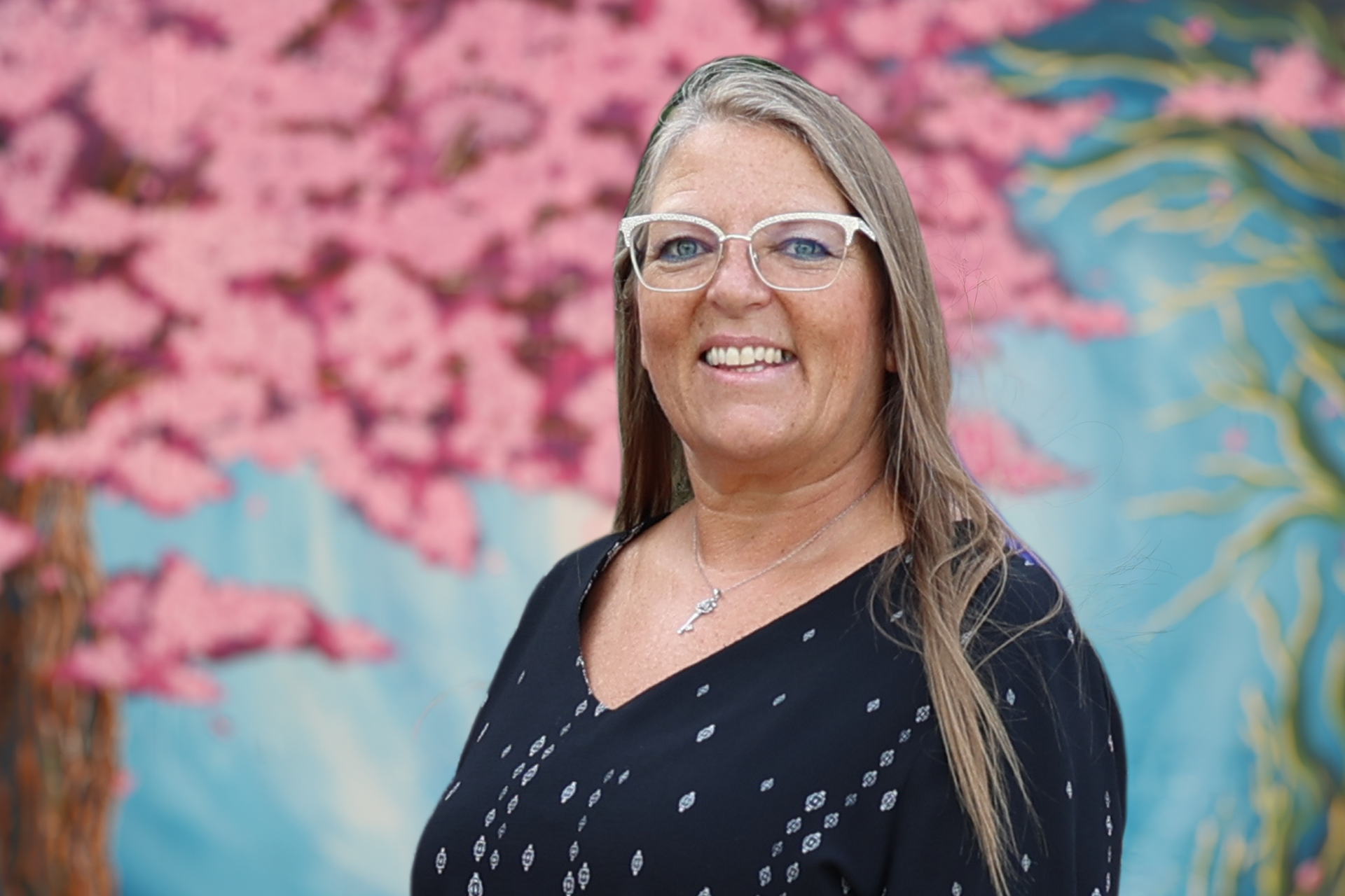 Smiling woman with glasses in a dark blouse, standing before a colorful mural background