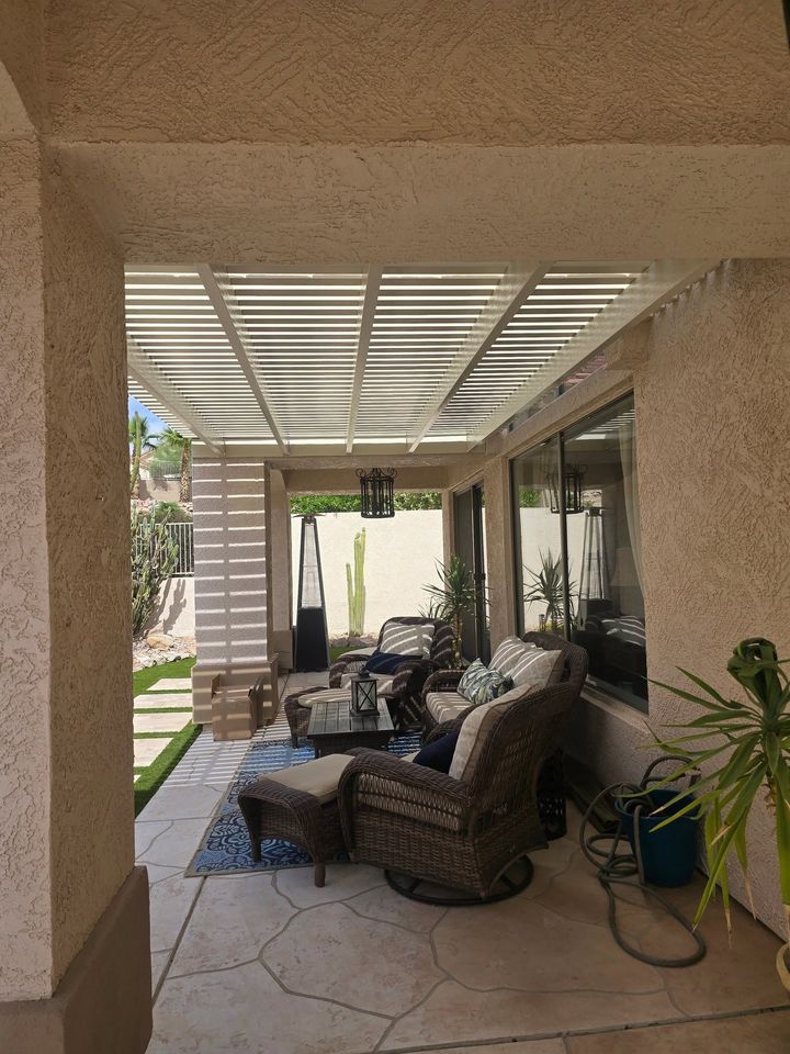Covered patio with wicker chairs, table, and potted plants beside a house and pool area.