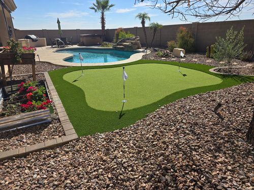 Backyard putting green beside a swimming pool, bordered by rocks and flower beds, under a sunny sky