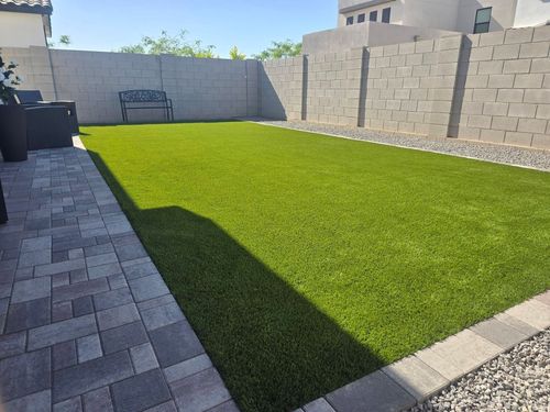 Backyard with green artificial turf, paver walkway, cinderblock walls, and a bench in the shade