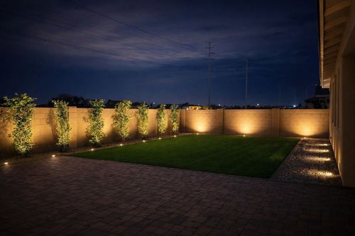 Illuminated backyard patio at dusk with stone walls, grass lawn, and pathway lights